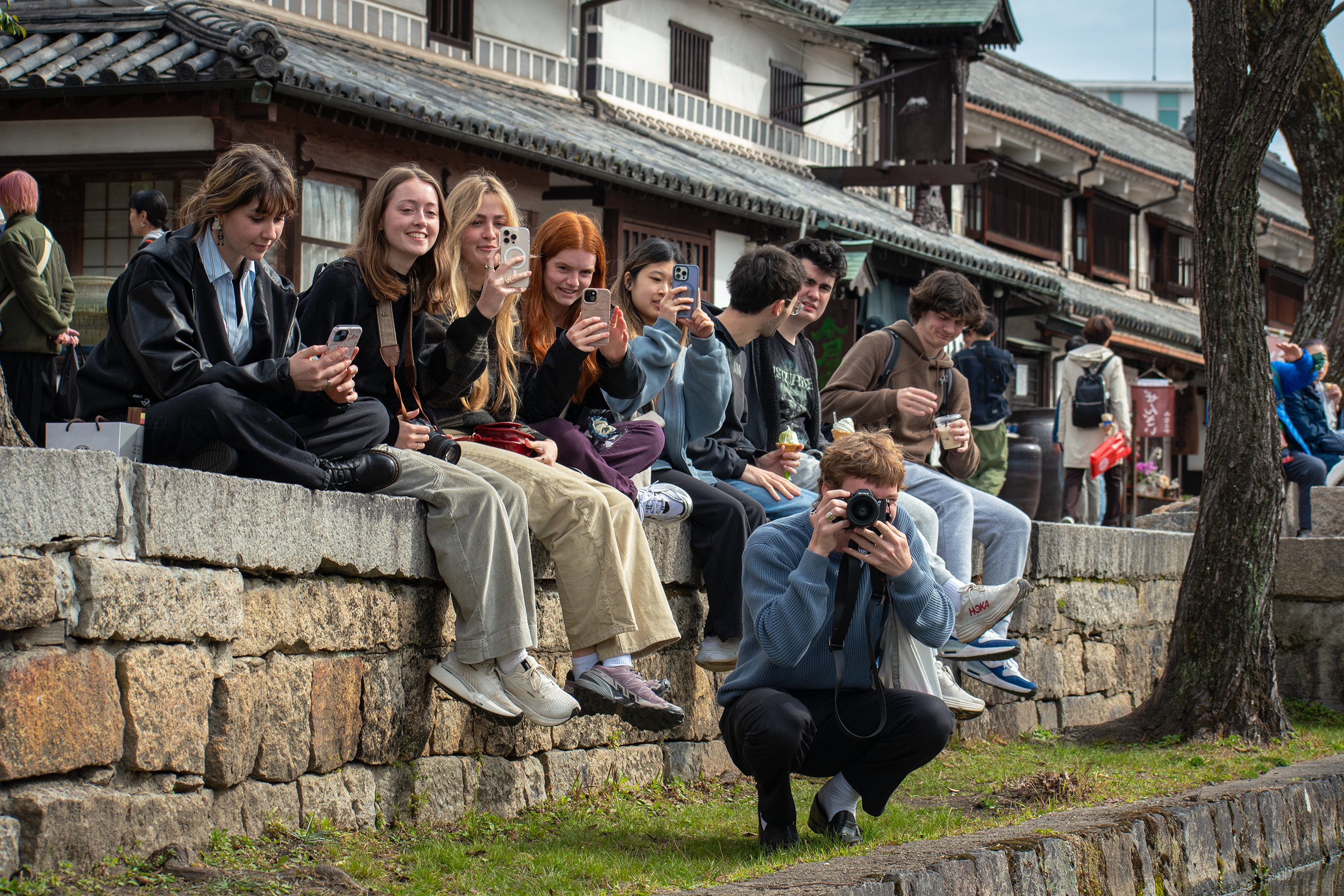 A group of young people sit on a stone wall, some smiling and using phones, while one person crouches in front taking a photo with a camera. Traditional buildings are in the background.