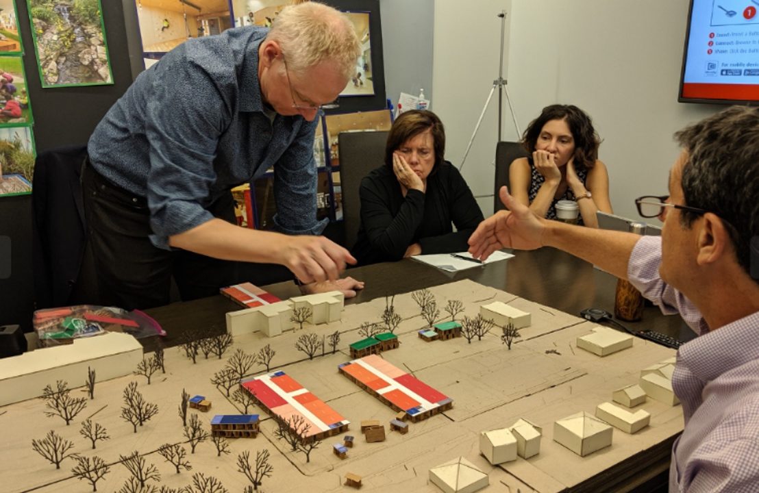 Four people discuss and gesture over an architectural model with small buildings, trees, vehicles, and printed images on a table in a meeting room.
