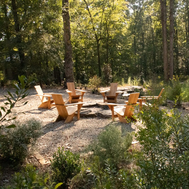 Five wooden chairs are arranged around a stone fire pit in a gravel clearing, surrounded by trees and green foliage in an outdoor setting.