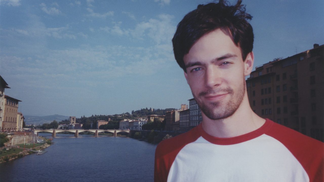 A man wearing a white and red shirt stands by a river with historic buildings and a bridge in the background under a blue sky.