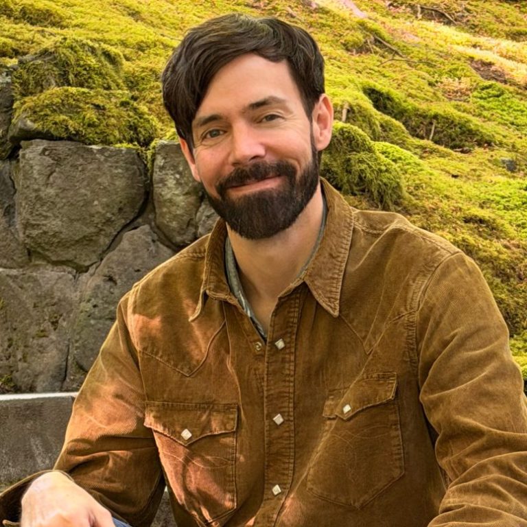 A man with a beard sits on stone steps surrounded by green moss and rocks in a lush outdoor setting.