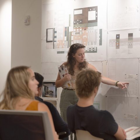 A woman stands and points to architectural plans on a wall while several people seated in chairs listen and watch.