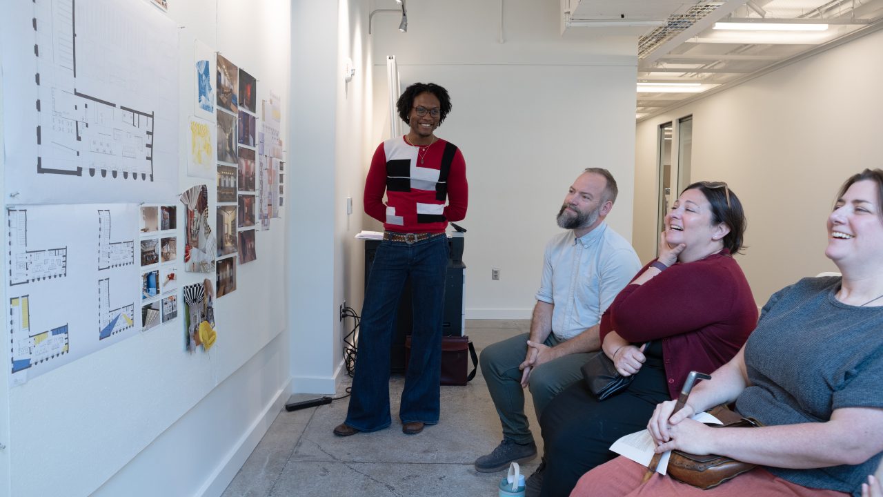 A person stands by a wall with floor plans and images, presenting to three seated people who are smiling in a bright, modern office space.