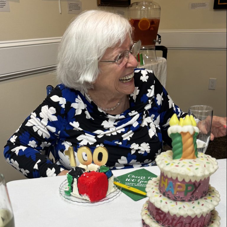 An elderly woman with white hair and glasses smiles at a table with a 100 candle, a fake cake, and champagne, celebrating her 100th birthday.