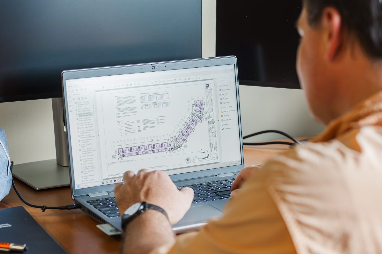 Person in a light orange shirt working on a laptop displaying a technical diagram at a desk with two monitors and office supplies.