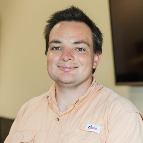 A man with short dark hair wearing a peach button-up shirt stands in a computer lab, smiling with hands clasped in front of him.