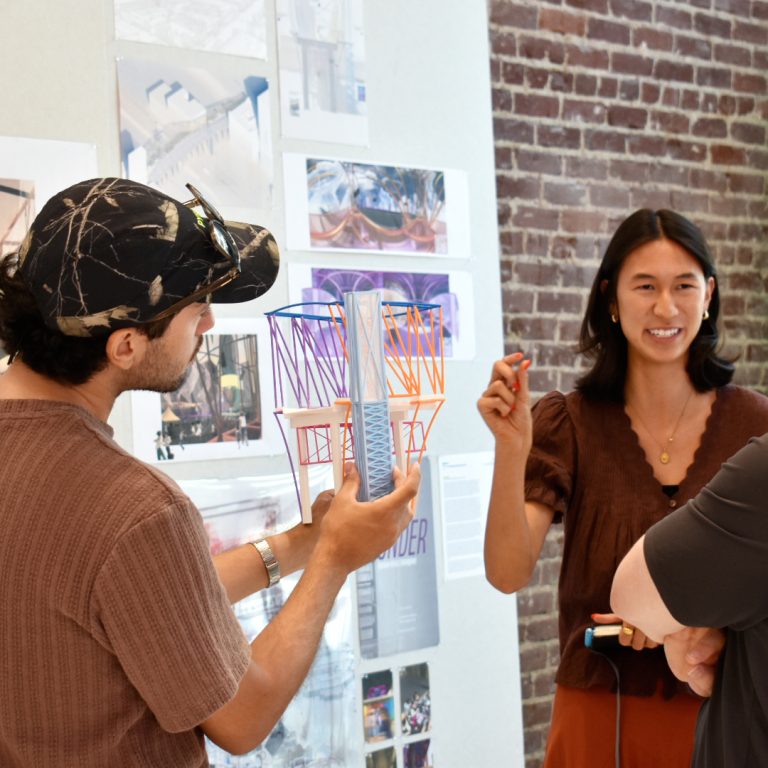 Three people discuss an architectural model and design plans displayed on a wall in a well-lit room with a brick wall and sketches.