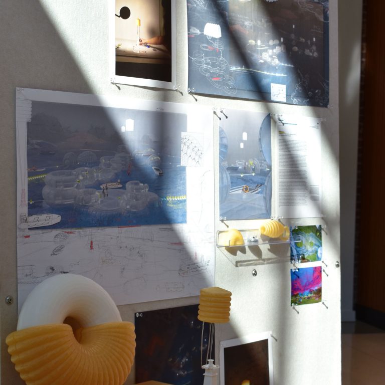 Design project display with 3D printed objects, architectural sketches, and concept images pinned to a wall, illuminated by sunlight. A desk holds more 3D prints, papers, and a remote control.