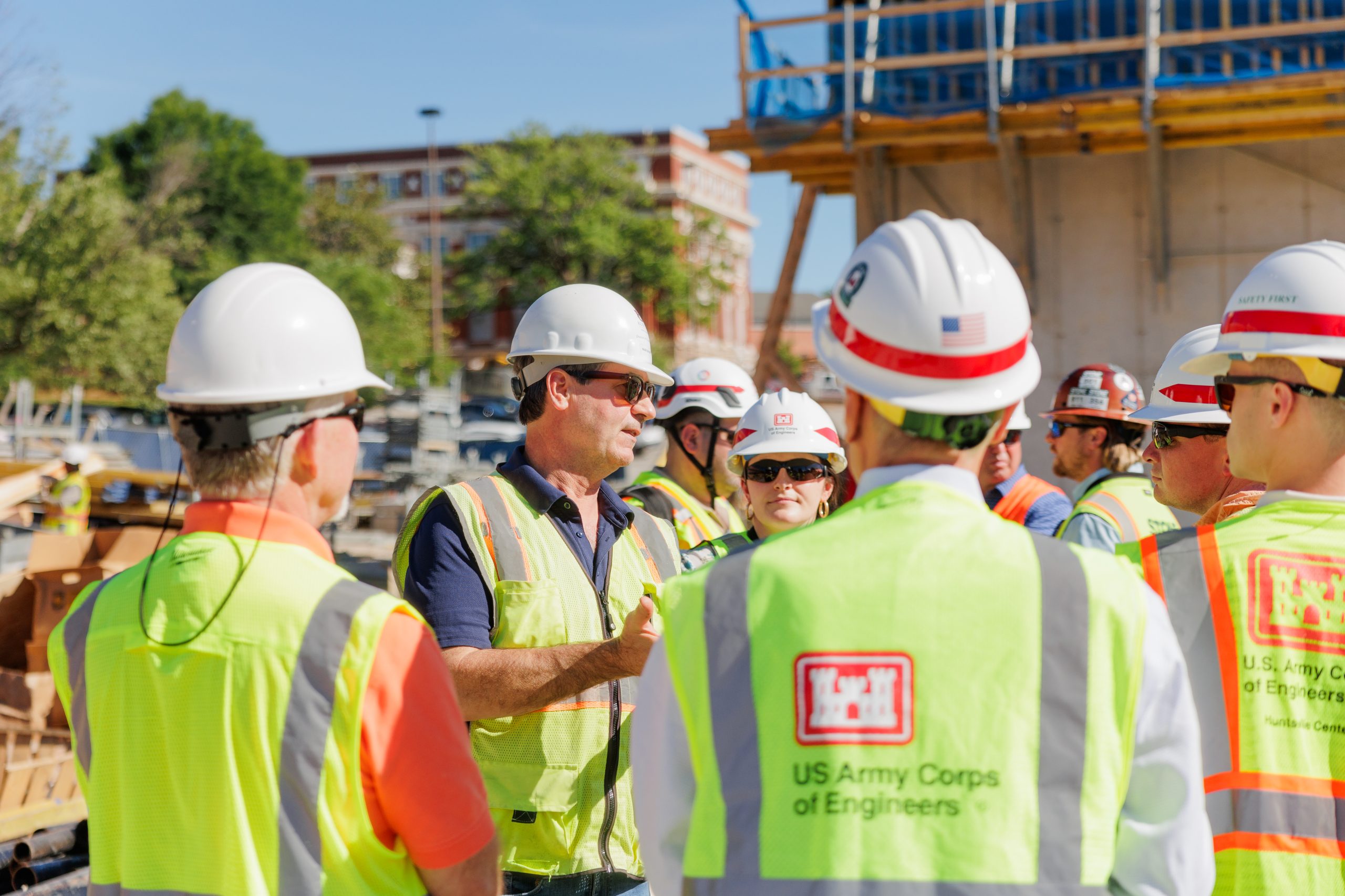 Group of people wearing hard hats and safety vests with US Army Corps of Engineers logos gathered at a construction site, listening to a discussion.