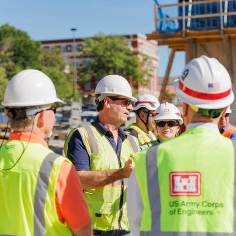 Group of people wearing hard hats and safety vests with US Army Corps of Engineers logos gathered at a construction site, listening to a discussion.