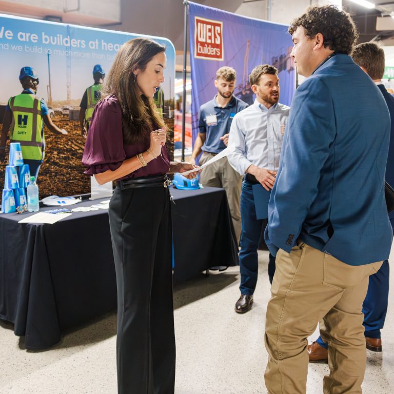 People converse at a career fair booth with construction banners in the background. The table displays brochures, hard hats, and bottles. Other attendees are visible nearby.