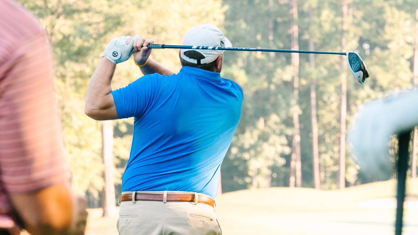 A man in a blue shirt and khaki shorts swings a golf club on a sunny golf course, with trees in the background.