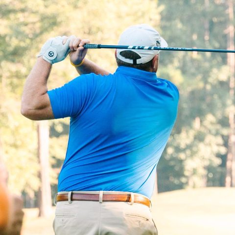 A man in a blue shirt and khaki shorts swings a golf club on a sunny golf course, with trees in the background.