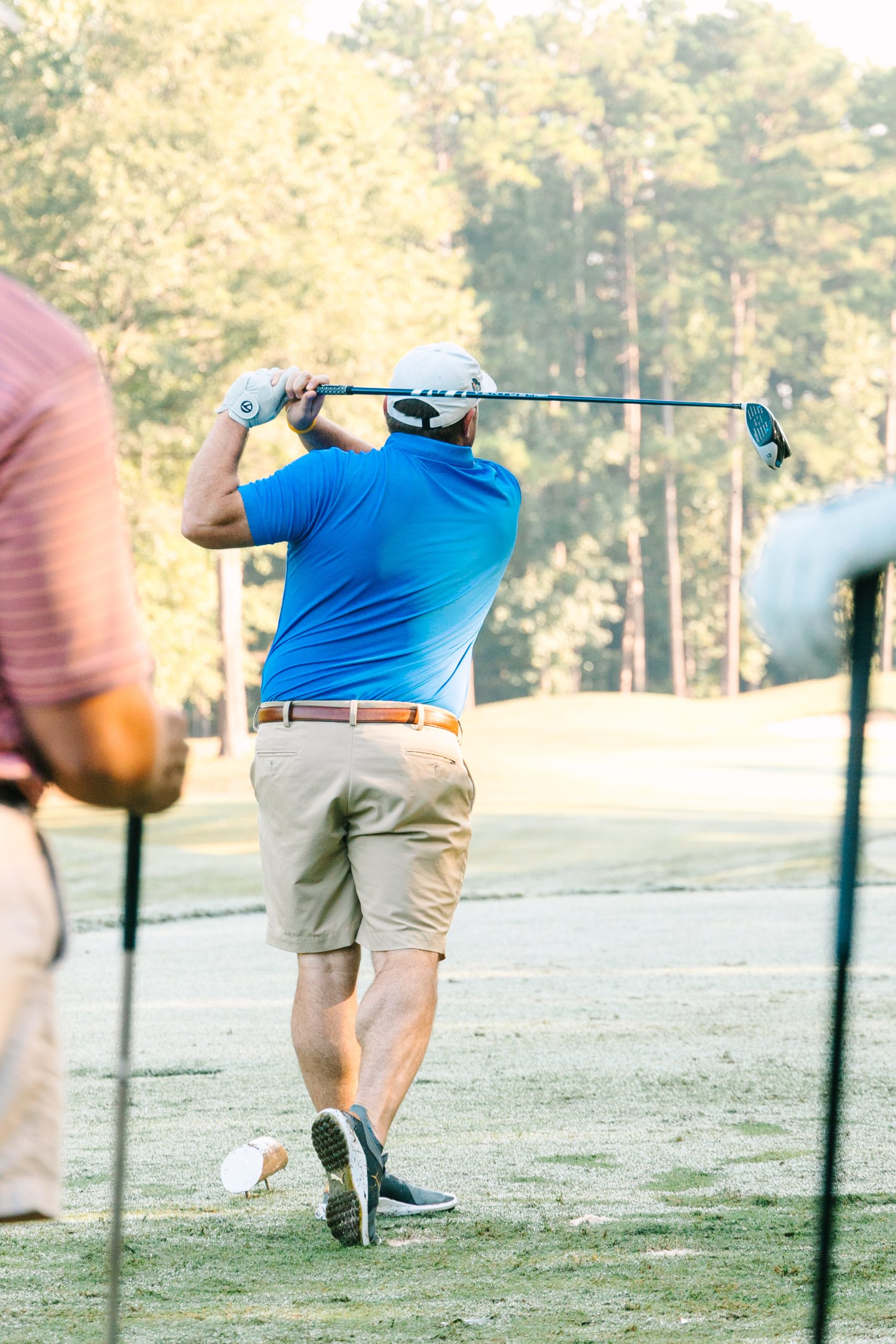 A man in a blue shirt and khaki shorts swings a golf club on a sunny golf course, with trees in the background.