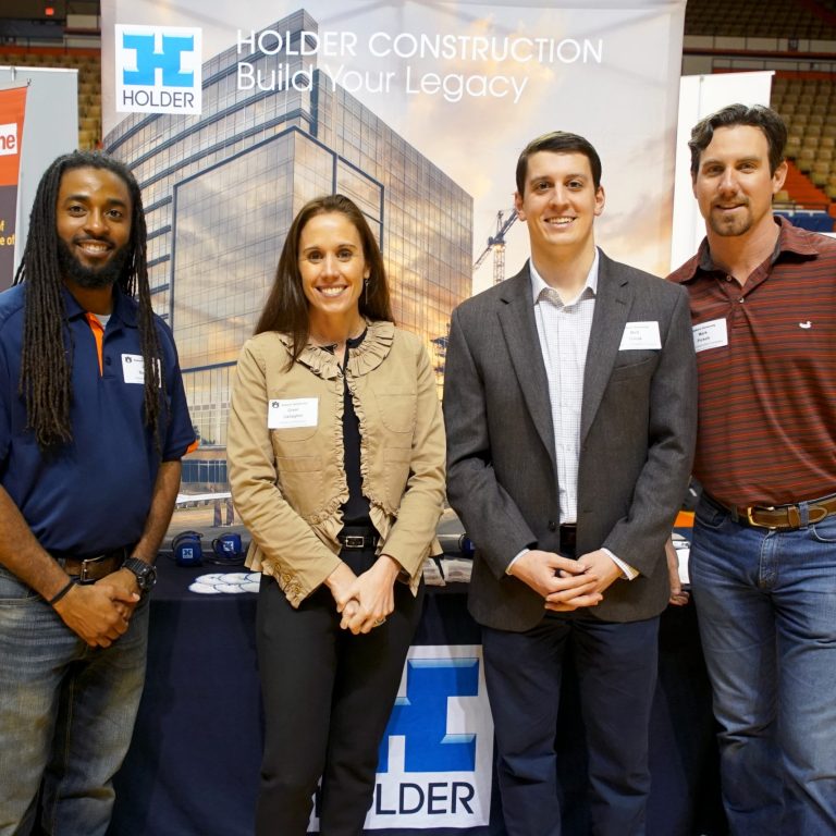 Four people stand in front of a Holder Construction booth at an indoor event, smiling at the camera.