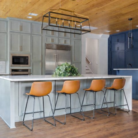 Modern kitchen with a large island, four brown barstools, green cabinets, navy accent wall, stainless appliances, and wood floors and ceiling.