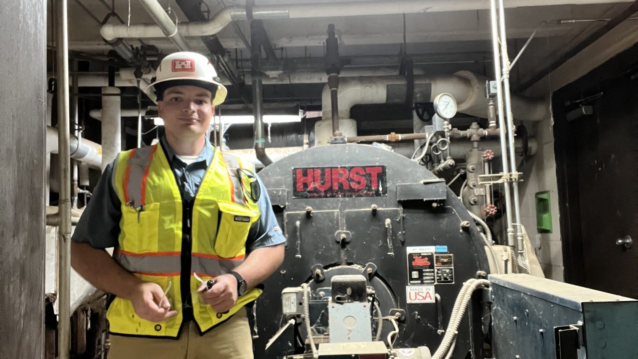 A person wearing safety gear stands in front of a large industrial Hurst boiler in a mechanical room with pipes and machinery visible.