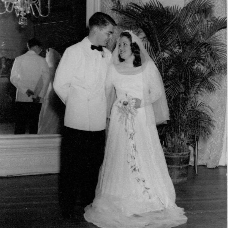 A bride in a long gown and veil stands next to a groom in a white tuxedo jacket; they smile at each other in a formal room with a chandelier and potted plant.