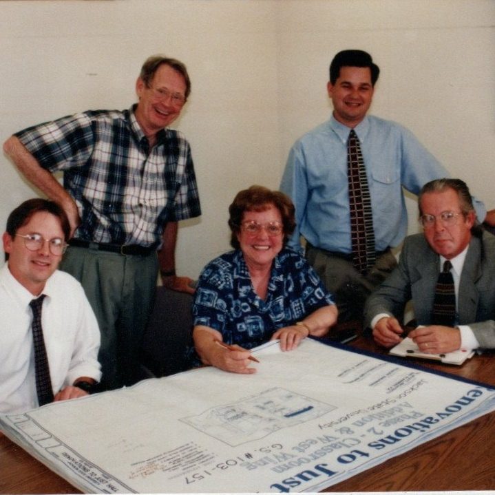 Five people, three men seated and two standing, gather around a table with documents and blueprints, appearing to discuss or review plans in a small office meeting room.