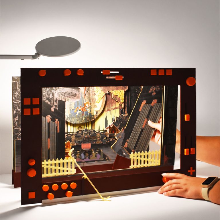A person sets up a tabletop diorama with cut-out figures depicting a stage scene, framed by a decorated border, near a lamp on a white surface.