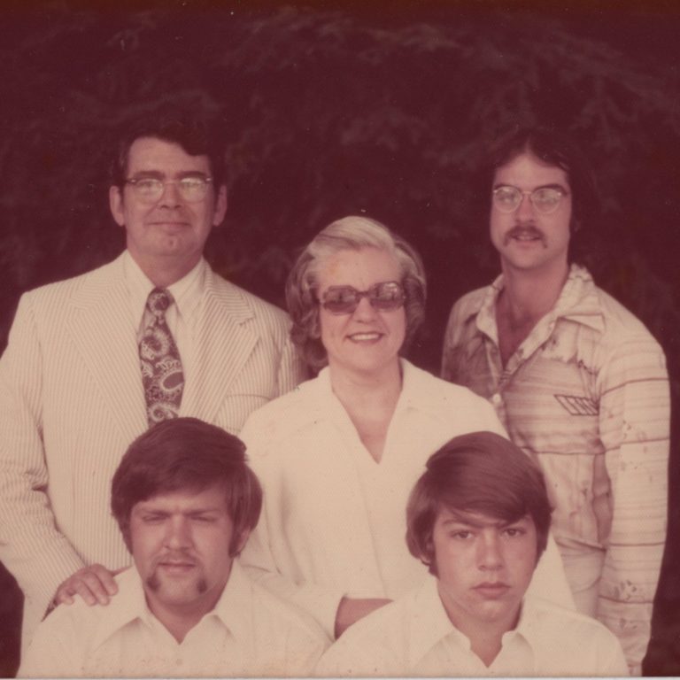 A group photo of two adult men, one adult woman, and two younger men, all wearing collared shirts, posed outdoors with trees in the background.
