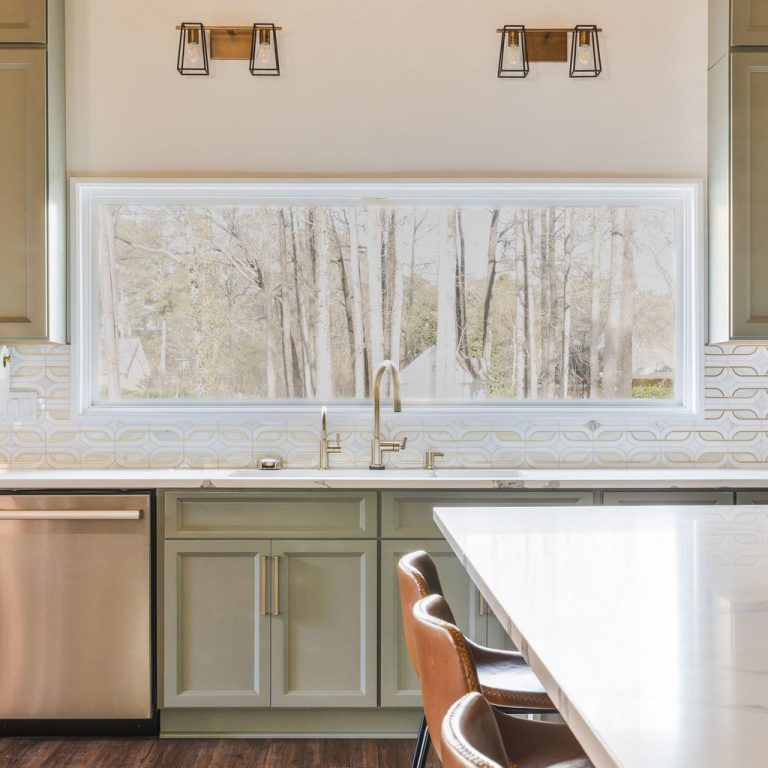Modern kitchen with green cabinets, white countertop, brown leather barstools, stainless steel appliances, and a large window above the sink showing trees outside.