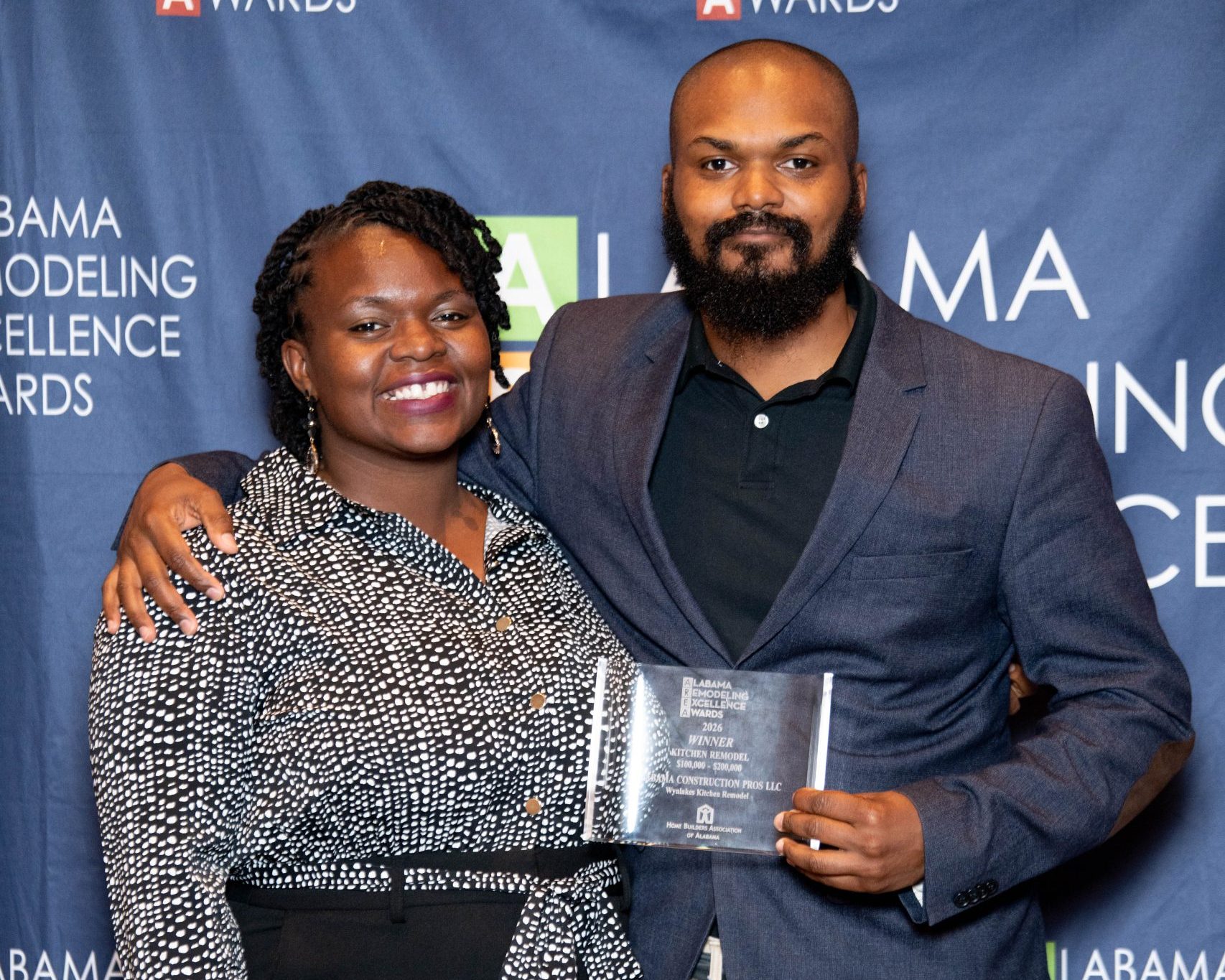 Two people stand together smiling at the Alabama Remodeling Excellence Awards; one holds a glass award plaque, both pose in front of an event backdrop.
