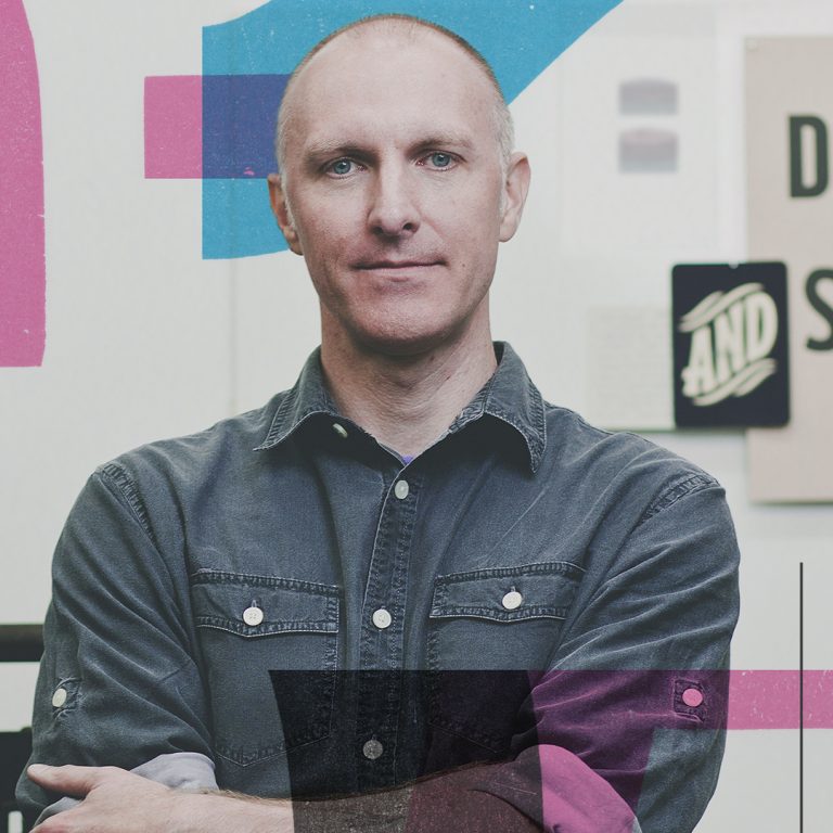 A man with short hair stands with arms crossed in front of a wall with posters, including one that says DESIGN IS SMART..