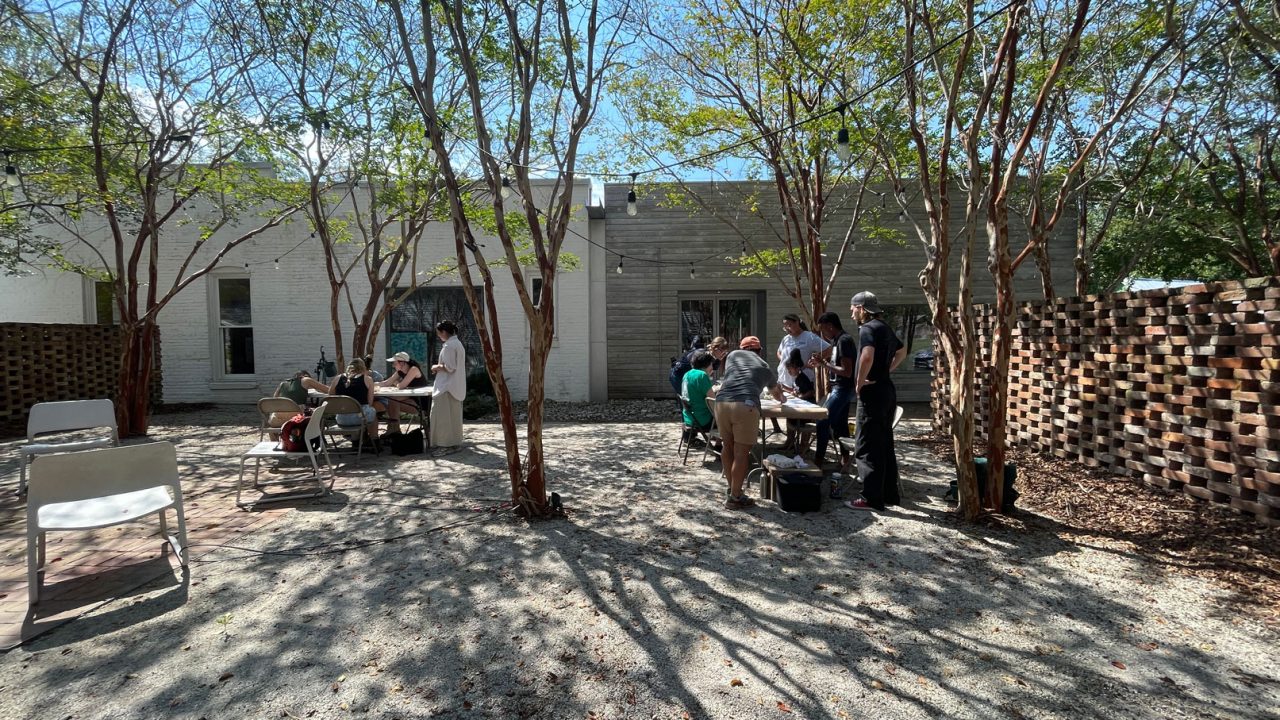 People sit and stand around tables in a sunlit outdoor courtyard with trees, gravel ground, and a brick building in the background.