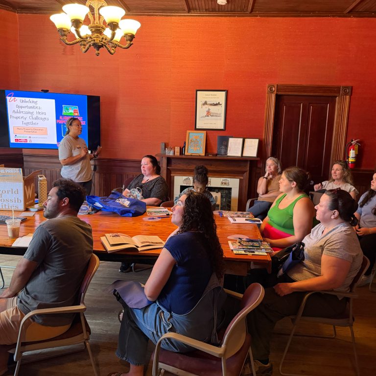 A group of people sit around a table listening to a speaker giving a presentation in a wood-paneled room with a TV screen displaying slides.