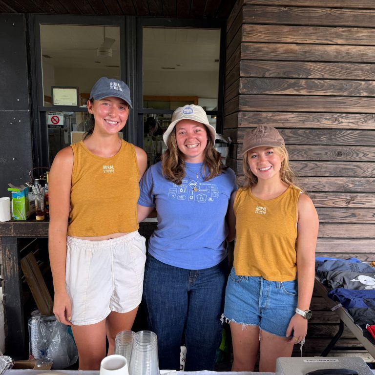 Three people stand side by side smiling behind a counter with cups and items, two wearing yellow tank tops and hats, the third in a blue shirt and hat, in front of a wooden wall.