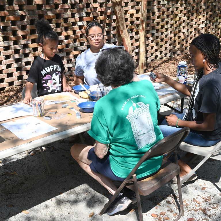 Four people sit at a table outdoors, working on arts and crafts projects with paint and paper supplies. A brick lattice wall is in the background.