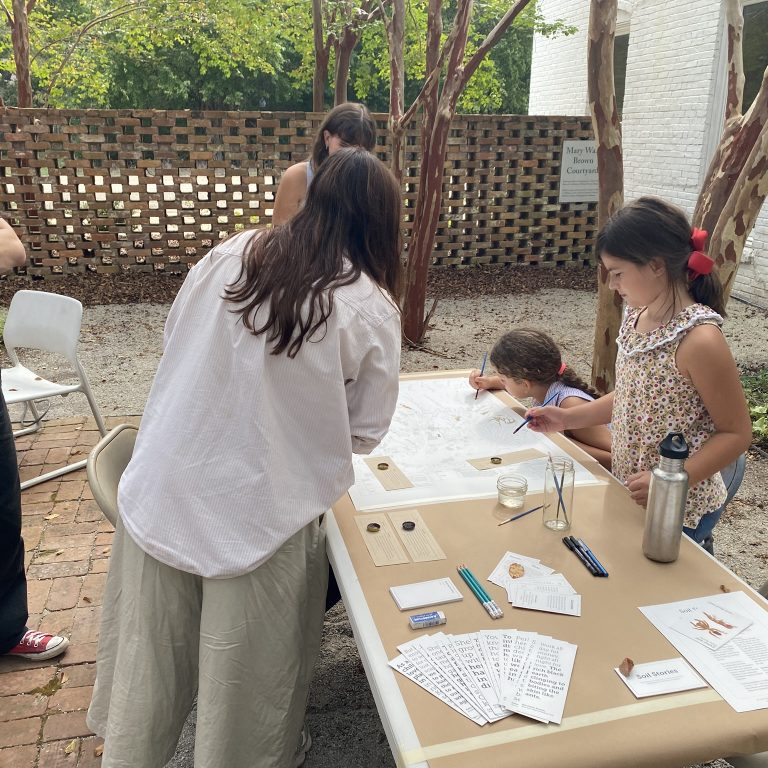Several people, including children, gather around a table outdoors, engaging in an activity with papers, pens, and informational materials. A man stands nearby with his arms crossed.