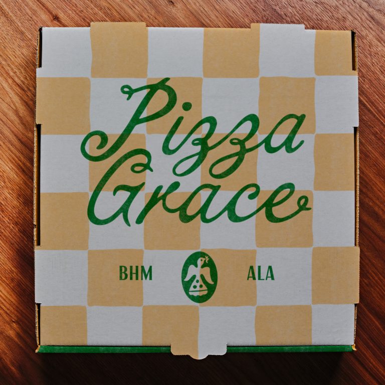 A Pizza Grace box with a yellow and white checker pattern sits on a wooden table, showing BHM and ALA beneath the logo.