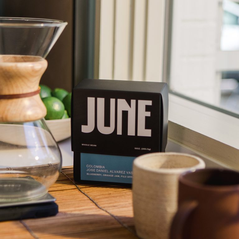 Box of June brand coffee next to a Chemex coffee maker, a mug, and a bowl of limes on a wooden surface by a window.