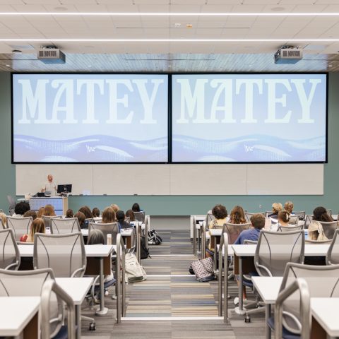 A classroom with students seated facing a lecturer at the front. Two large screens display the word MATEY in capital letters.