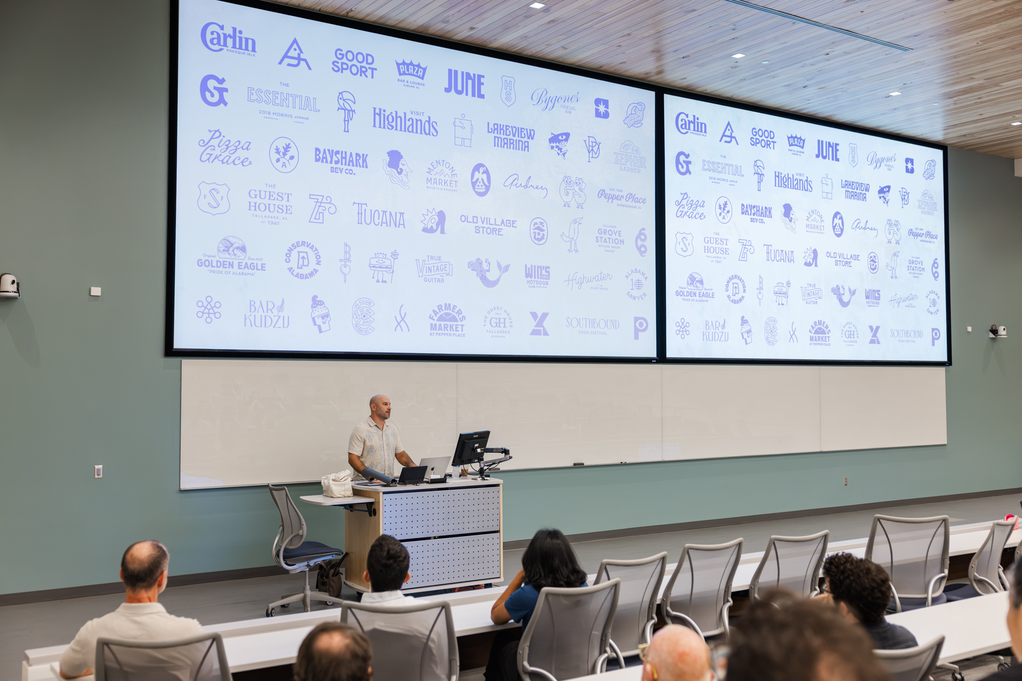 A person presents in a lecture hall with a slide showing dozens of business logos to an audience seated in rows.