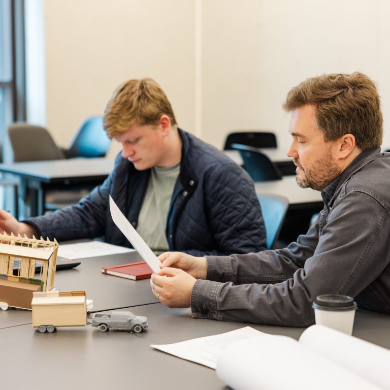 Two men sit at a table with papers, a laptop, and small architectural models, appearing focused on their work in a classroom setting.