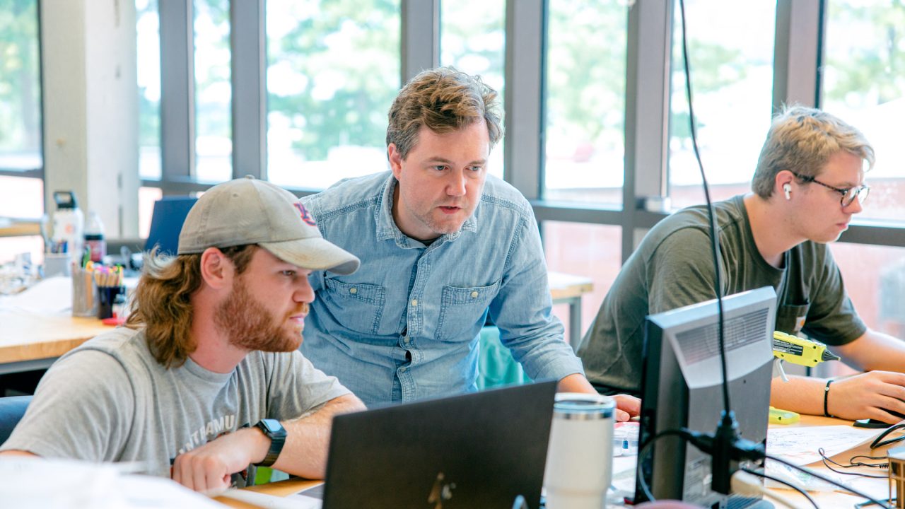 Three men work at desks with laptops. One man stands and looks at a screen while two others are seated and focused on their computers. Large windows are in the background.