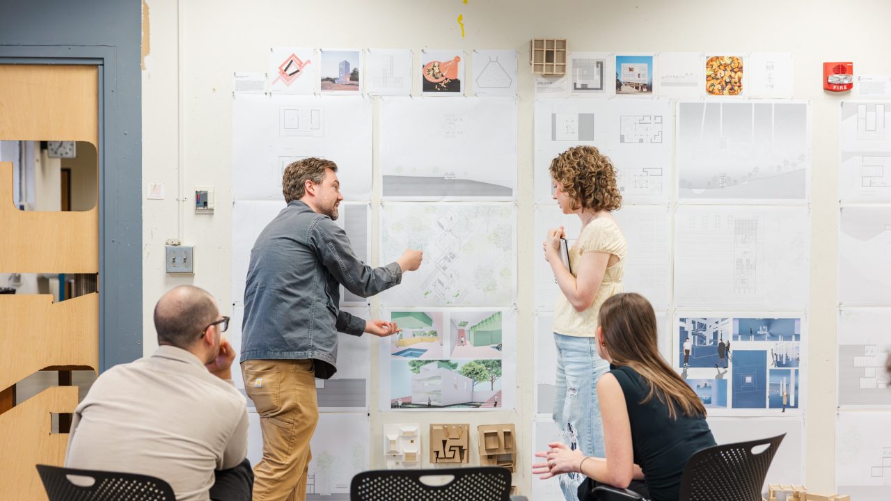 Four people in a classroom discuss architectural drawings and models displayed on a wall. Two stand and gesture toward the plans while two sit and observe.