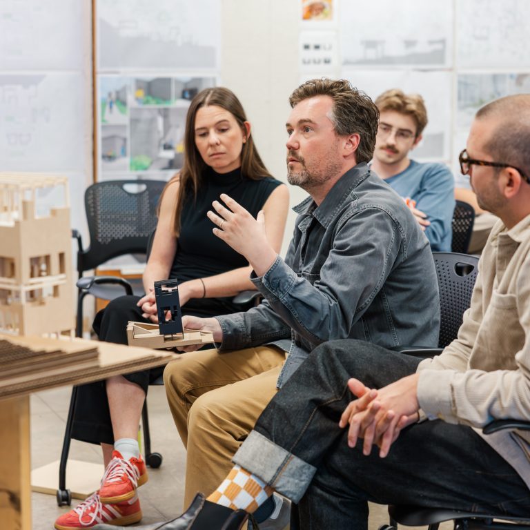 A man speaks while seated in a classroom, gesturing with his hand, as three other adults listen. Architectural drawings and a model are visible in the background.