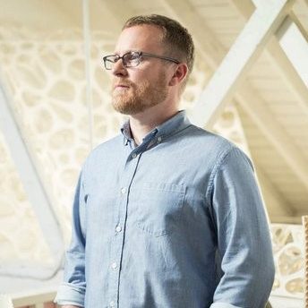 A man with glasses and a beard stands indoors with hands in pockets, wearing a light blue button-up shirt and jeans. The room has white walls and natural light.