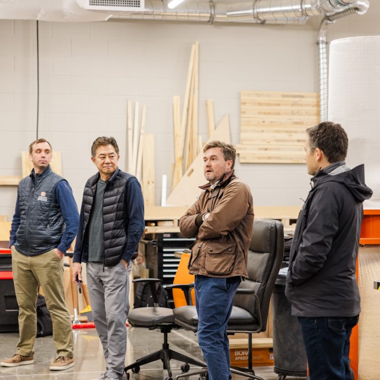 Four men stand and converse in a woodworking shop, surrounded by tools, machinery, and stacks of wood.