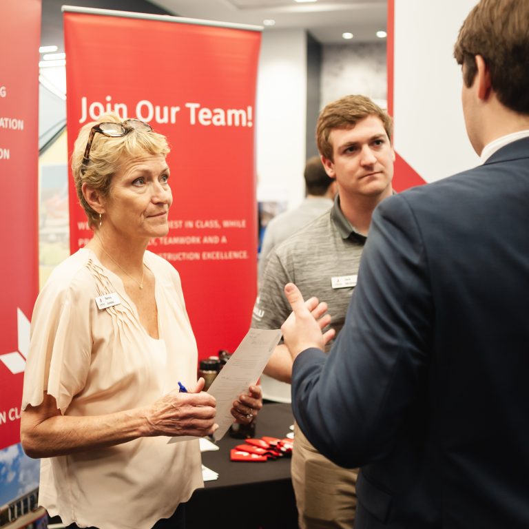 Three people talk at a career fair booth with Join Our Team! on a red banner in the background. One woman holds a clipboard while the two men listen and engage in conversation.