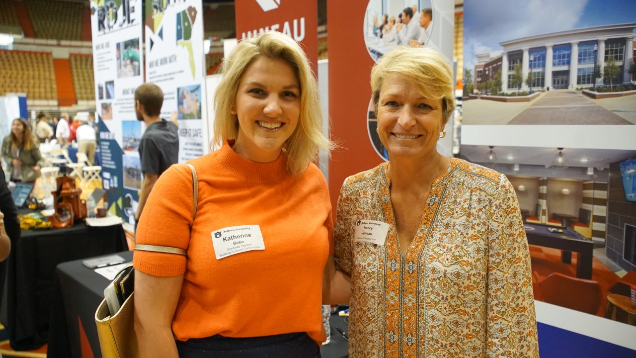 Two women wearing name tags stand side by side and smile at a business event with informational booths and displays in the background.