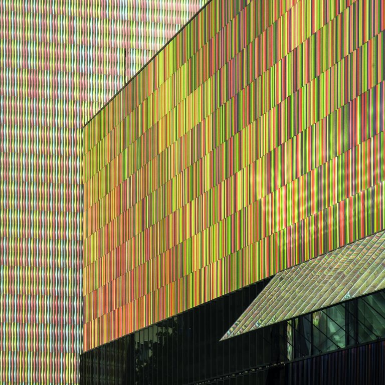 Modern building with exterior walls featuring a grid of colorful vertical stripes, one window, and a slanted glass section near the bottom right.