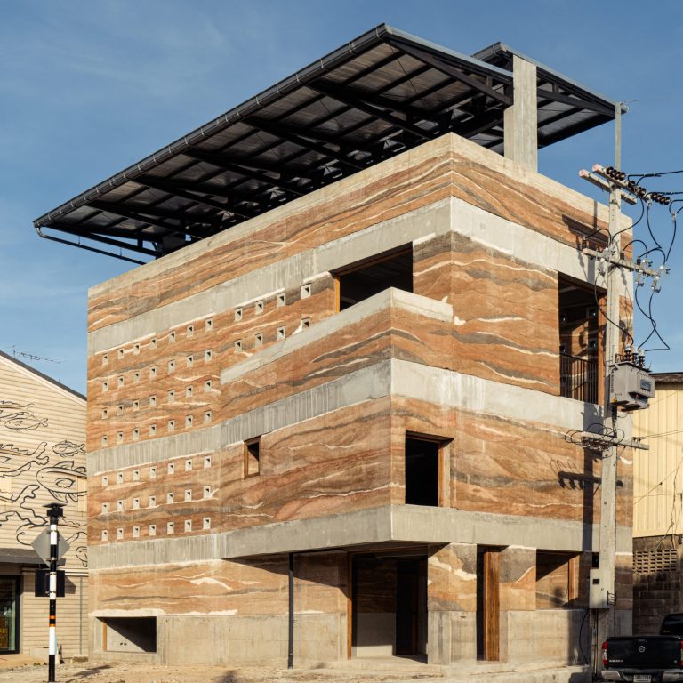 Four-story modern building with layered concrete and stone facade, small square windows, and a large solar panel roof structure, situated on a street corner.
