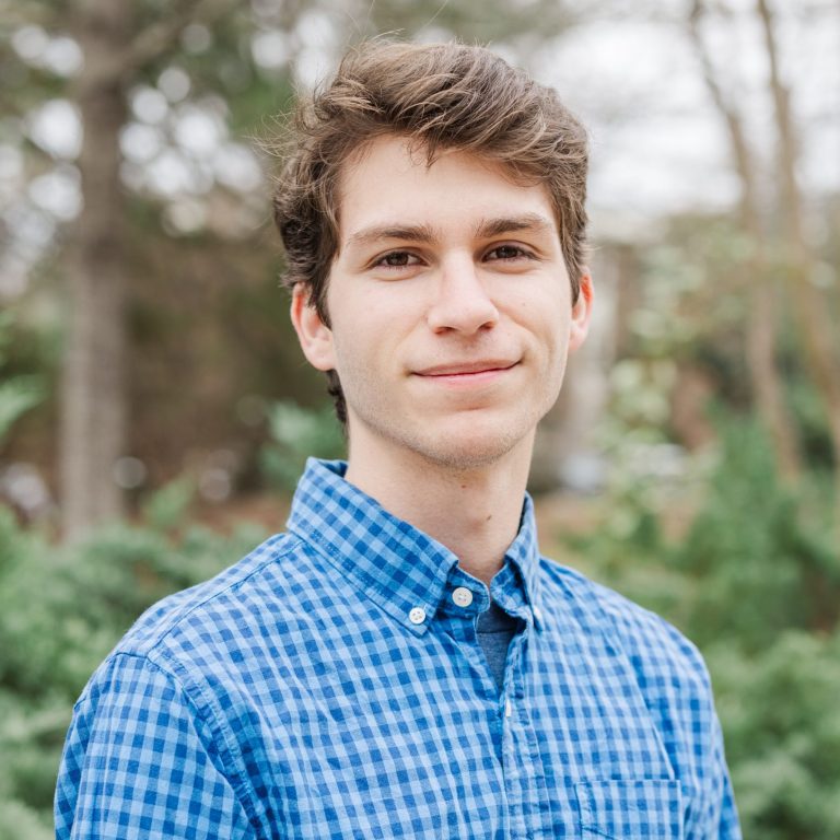 A young man with brown hair wearing a blue plaid shirt stands outdoors in front of green foliage, looking at the camera with a neutral expression.