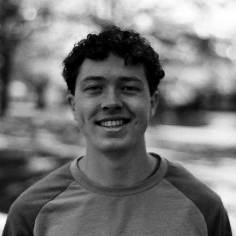 A young man with curly hair and a light-colored sweatshirt smiles outdoors, with trees and sunlight blurred in the background.