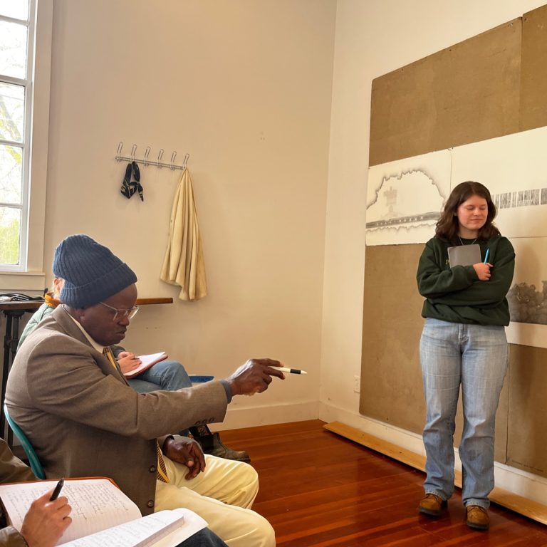 A woman stands by architectural drawings on a wall while others sit nearby, one man pointing and holding a notebook.